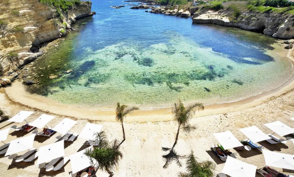 Spiaggia di sabbia con lettini e ombrelloni bianchi affacciata su una baia dalle acque cristalline