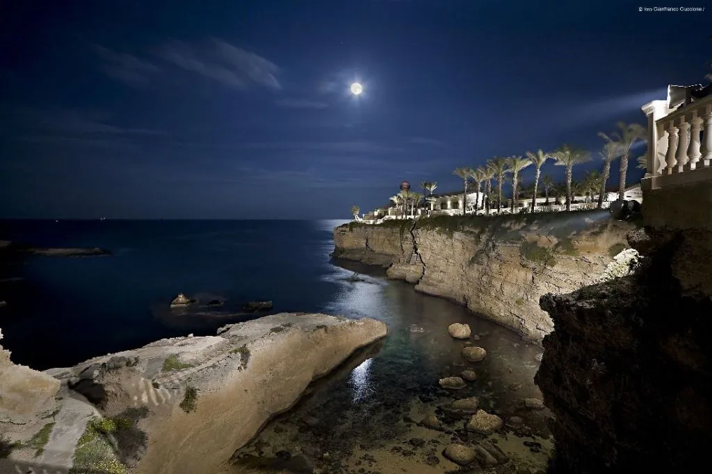 Vista notturna della costa rocciosa illuminata dalla luna piena con palme del resort
