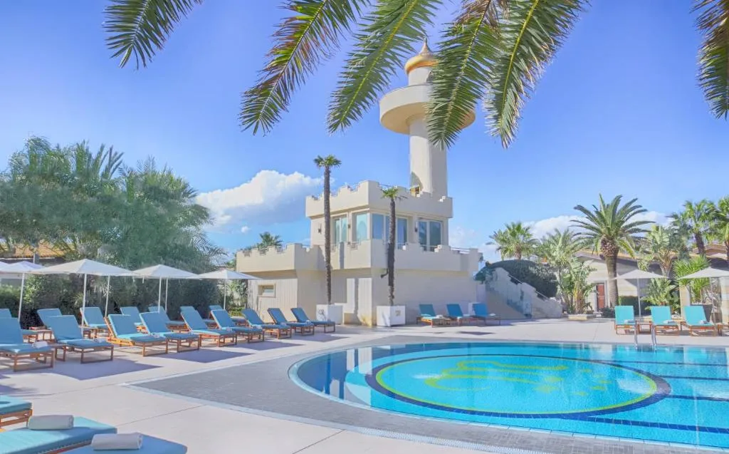 Piscina esterna con lettini blu, ombrelloni e torre in stile moresco tra le palme