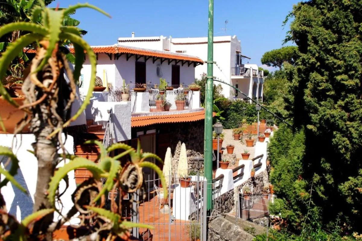 Esterno dell'hotel con edifici bianchi, balconi fioriti con piante in vaso e vegetazione