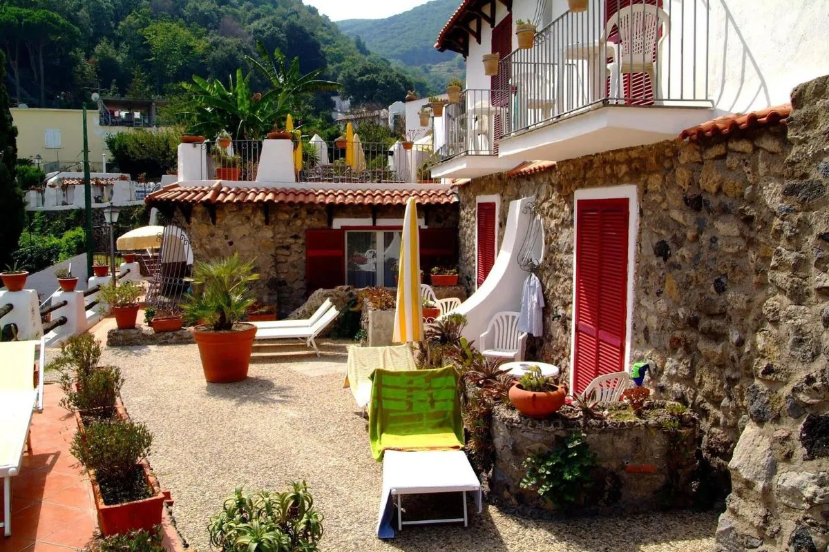 Cortile esterno in pietra con lettini, ombrelloni, piante in vaso e persiane rosse
