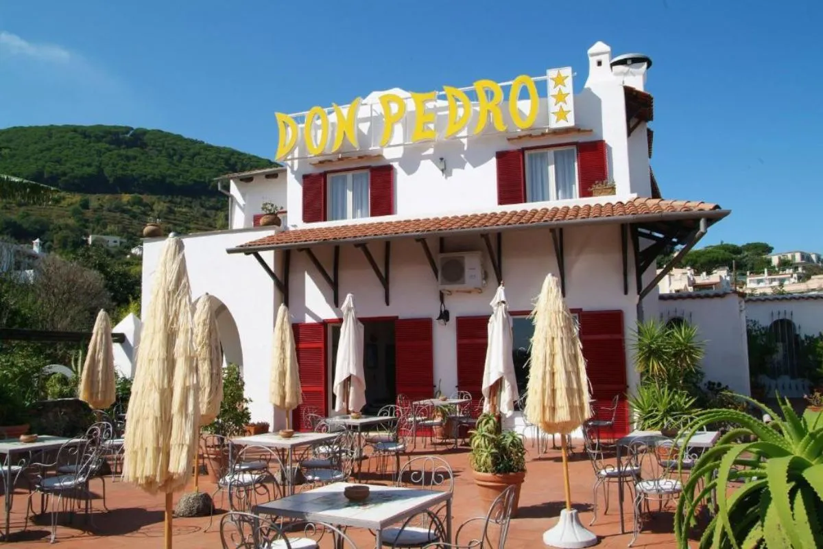 Facciata dell'Hotel Don Pedro con insegna gialla, persiane rosse e terrazza con tavoli e ombrelloni