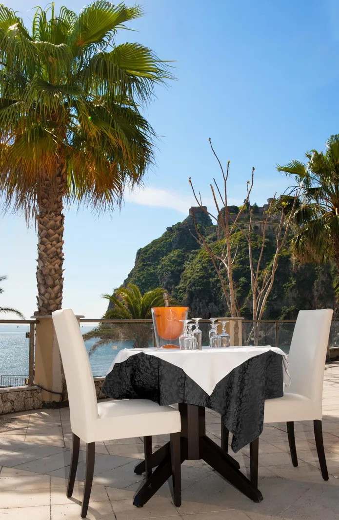 Tavolo su terrazza con vista sul mare, palme e promontorio roccioso