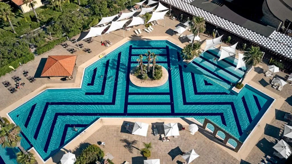 Piscina dal design geometrico con isola di palme centrale e ombrelloni bianchi vista dall'alto