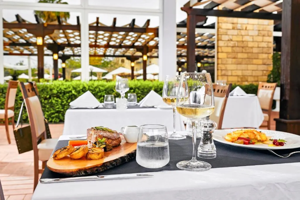 Tavolo del ristorante all'aperto con piatti di carne e calice di vino bianco sotto pergolato