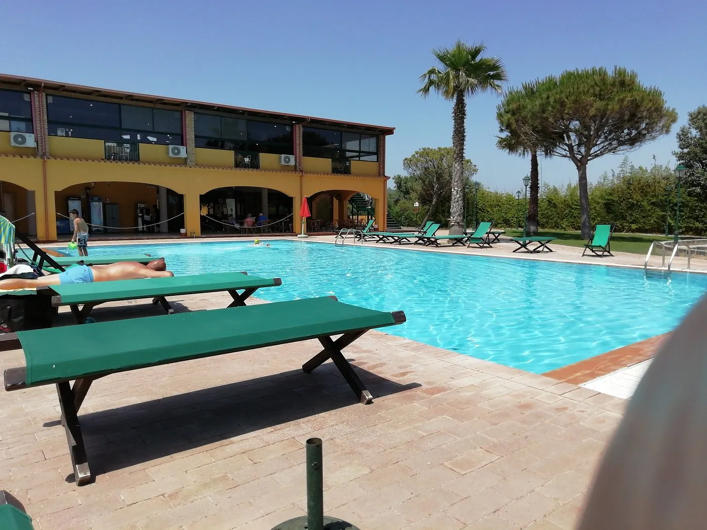 Piscina con lettini verdi e edificio giallo dell'hotel con ampie vetrate