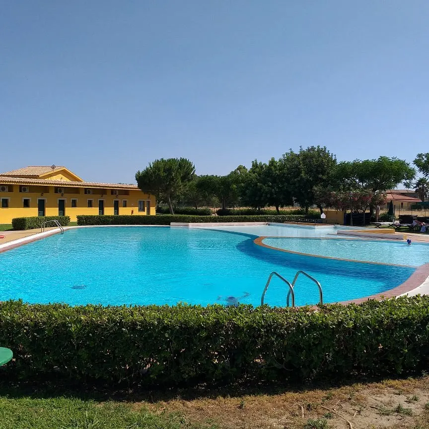 Piscina dalla forma curva con siepe verde e edificio giallo dell'hotel sullo sfondo