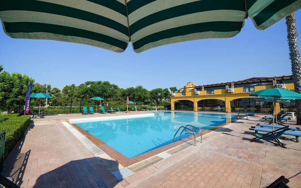 Piscina vista da sotto un ombrellone a righe verdi con edificio giallo dell'hotel
