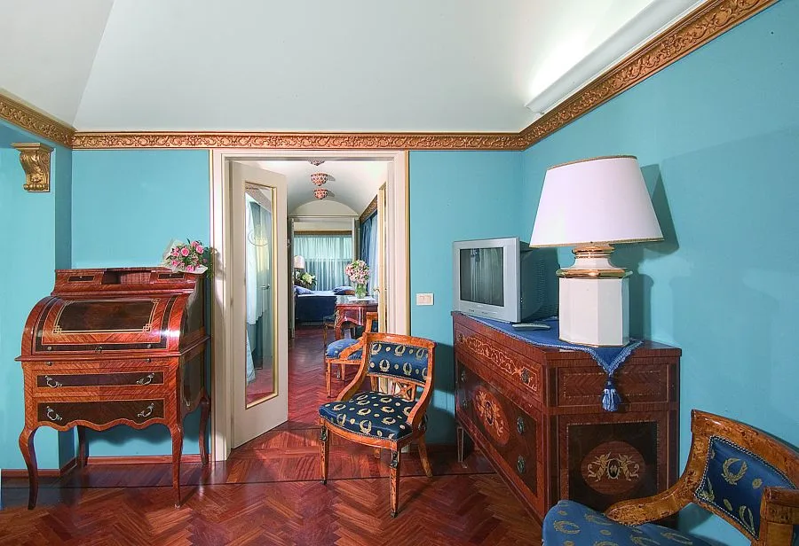 Soggiorno di camera con pareti azzurre, mobili in legno intarsiato e vista sulla camera da letto