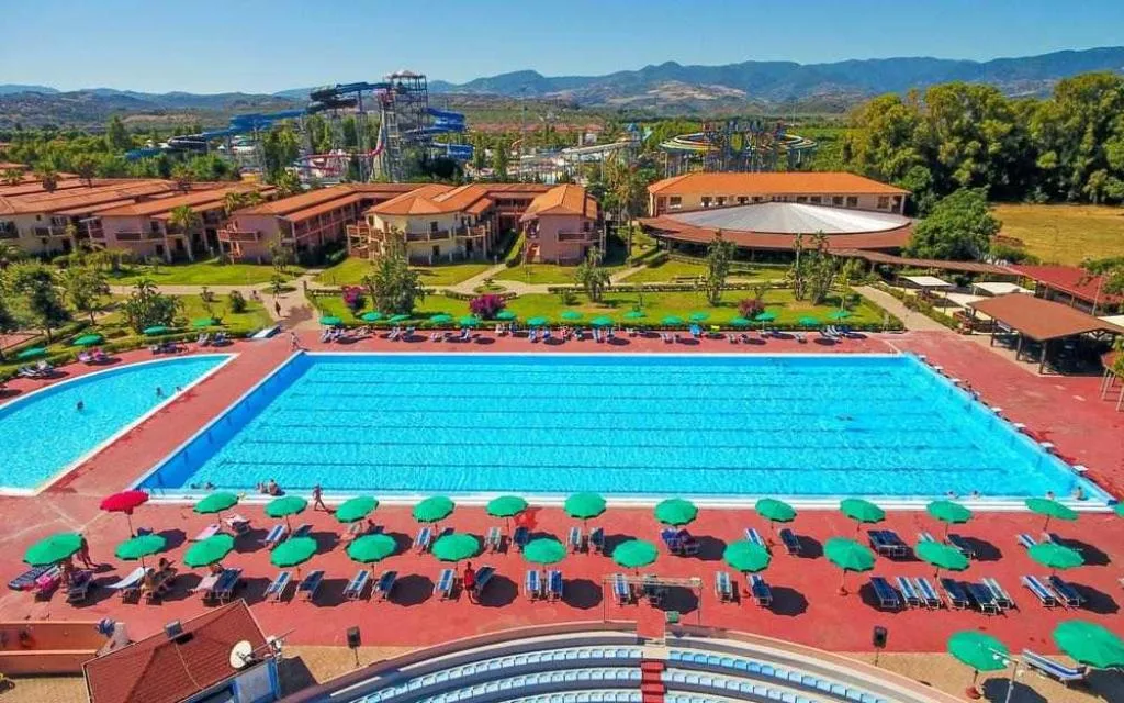 Grande piscina del resort con ombrelloni verdi, lettini e parco acquatico sullo sfondo tra le montagne