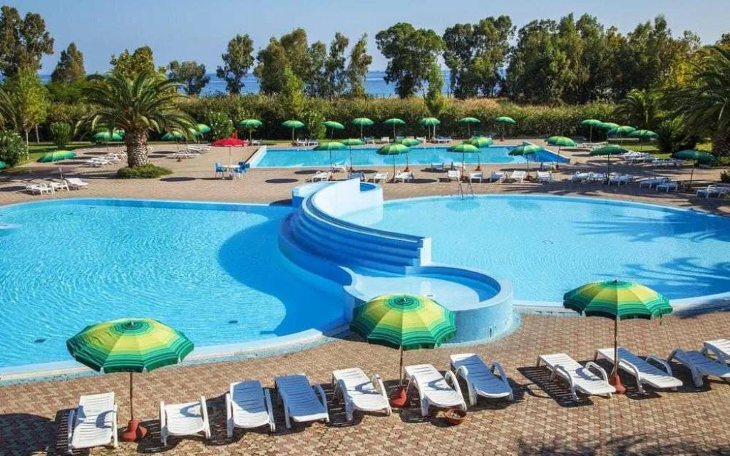 Piscina dalle forme curve con ombrelloni verdi e gialli, lettini bianchi e mare sullo sfondo
