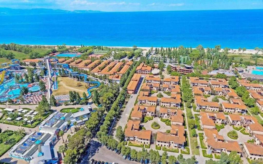 Panoramica aerea del resort fronte mare con edifici dai tetti rossi, parco acquatico e spiaggia