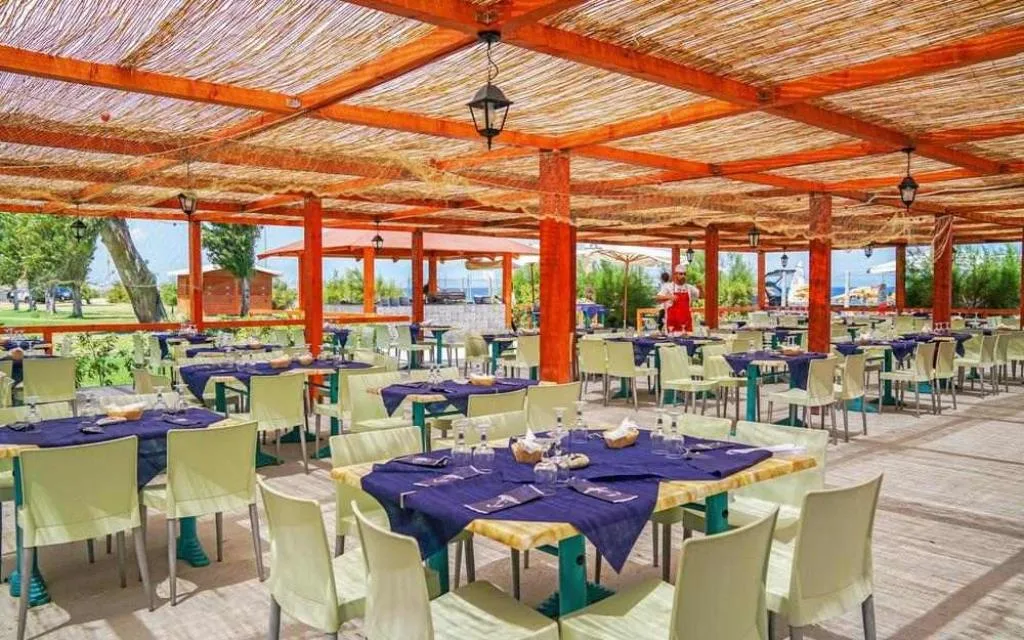 Ristorante all'aperto con pergolato in canne, tavoli apparecchiati e vista sul giardino verso il mare