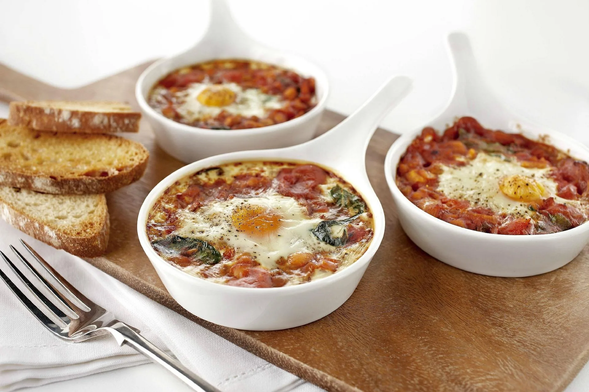 Uova al forno con salsa di pomodoro e spinaci in pirofile bianche con pane tostato
