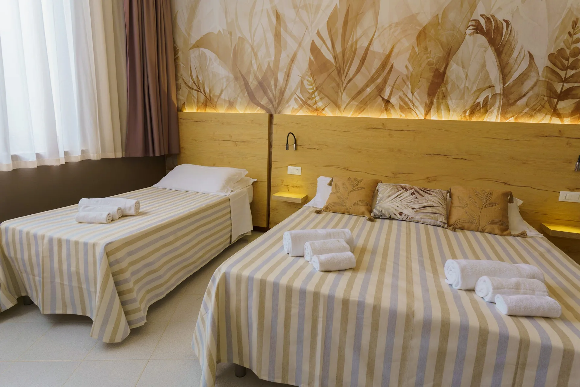 Camera con letto matrimoniale e letto singolo, testiera in legno e parete decorata con motivo botanico