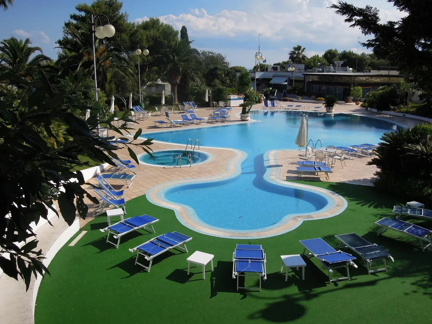 Piscina panoramica con prato verde, lettini blu e ombrelloni immersa in un giardino di palme