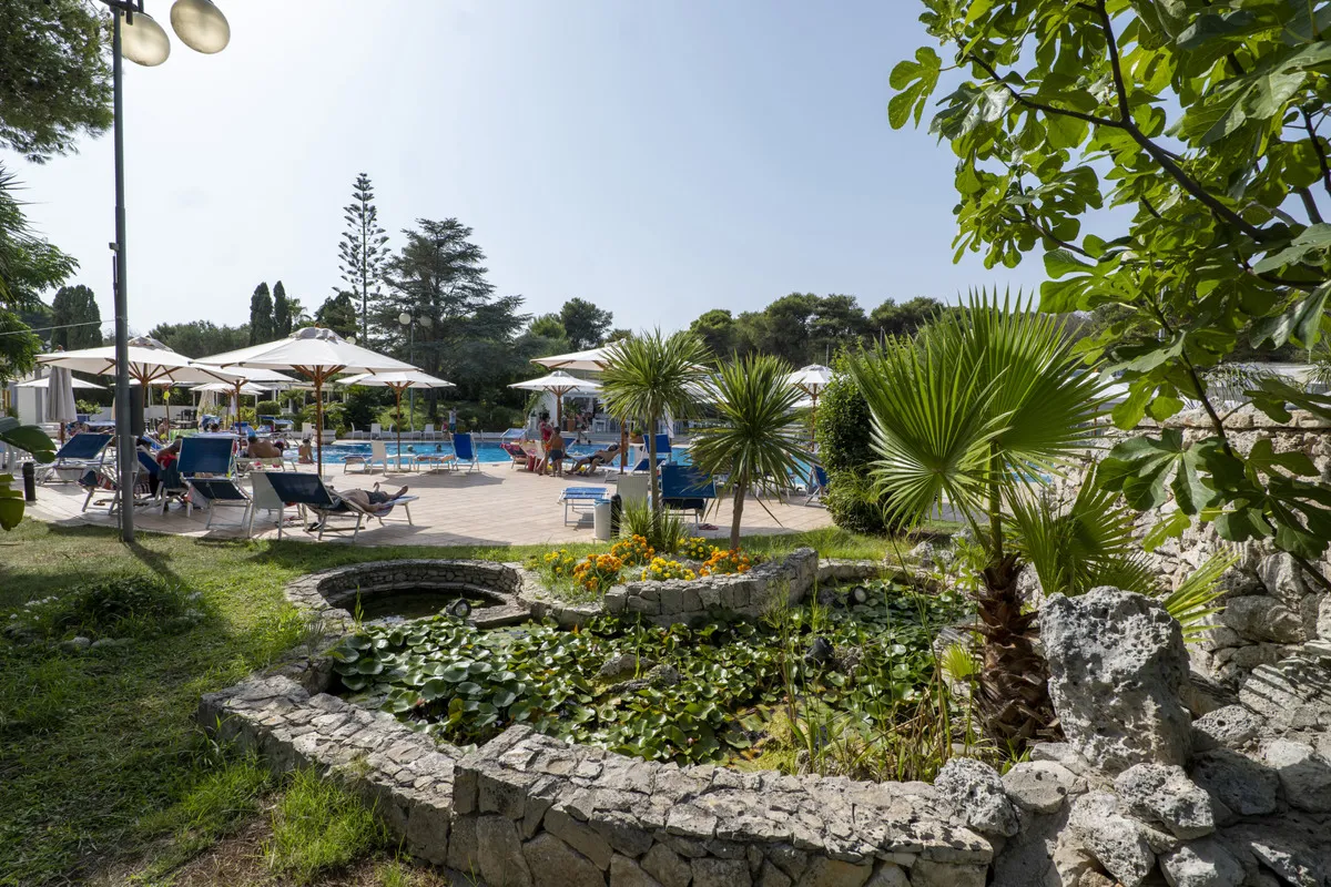Giardino con laghetto in pietra e piante acquatiche affacciato sulla zona piscina con ombrelloni bianchi