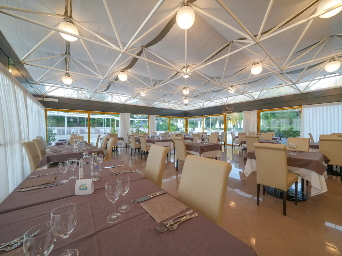 Interno del ristorante con tavoli eleganti, sedie beige e grandi finestre affacciate sul giardino