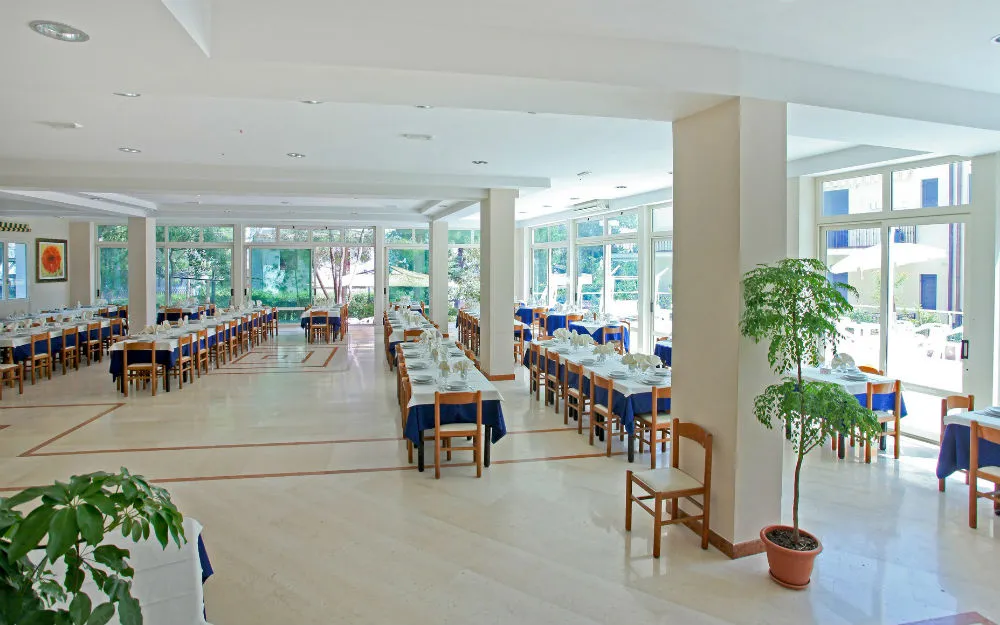Ampia sala da pranzo luminosa con tavoli e tovaglie blu, sedie in legno e grandi vetrate