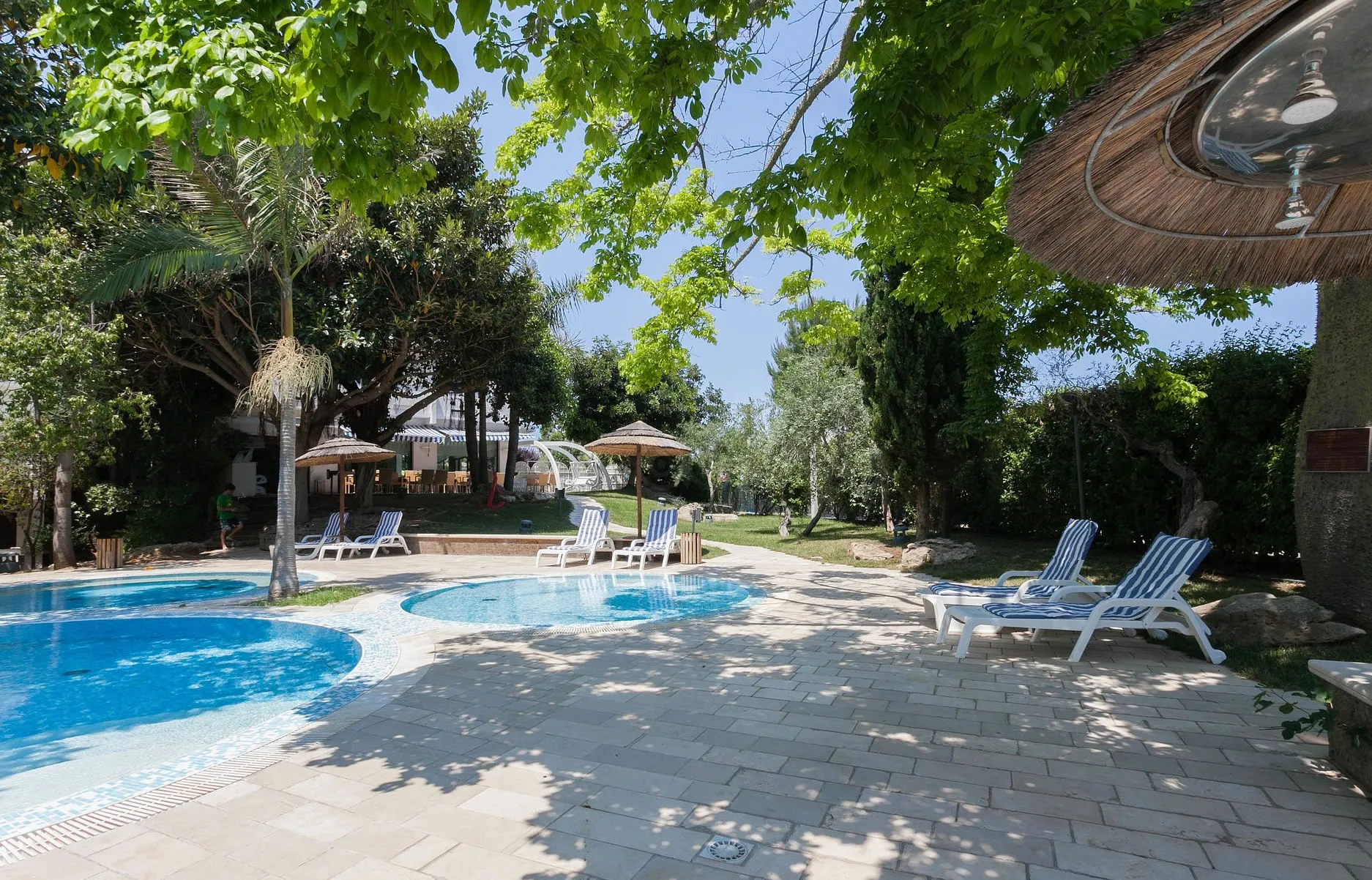 Area piscina con lettini a righe blu, ombrelloni in paglia e alberi ombrosi
