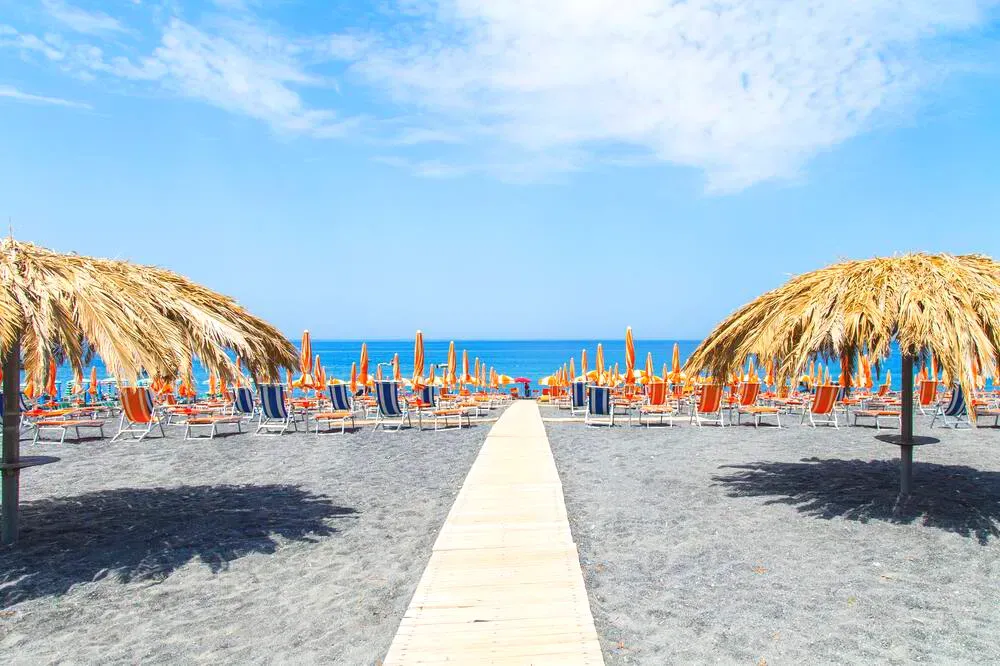 Spiaggia di sabbia scura con ombrelloni di paglia e lettini sul mare