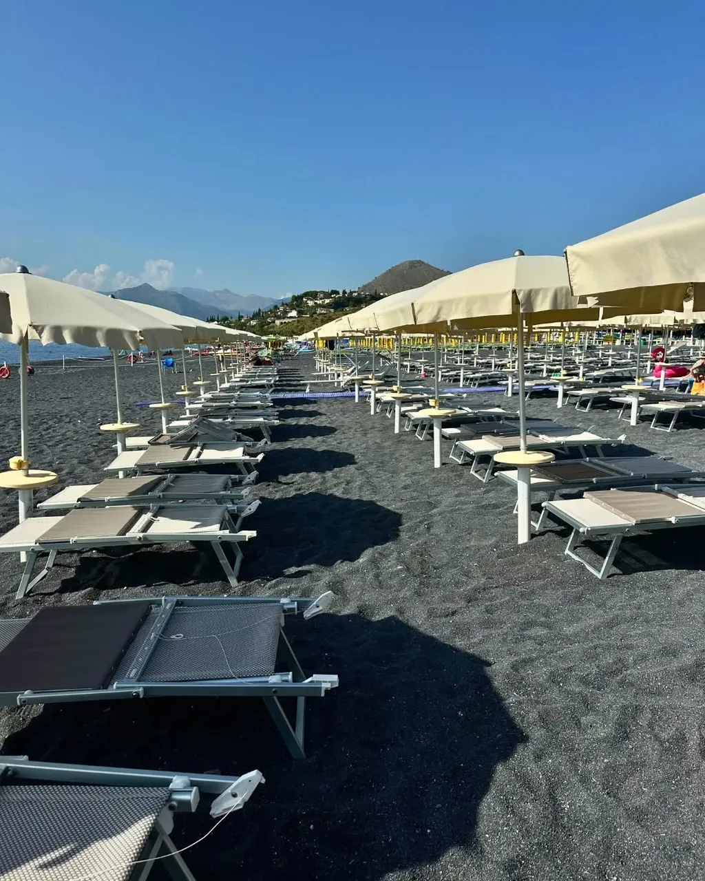 Lettini e ombrelloni in fila sulla spiaggia di sabbia nera con vista colline
