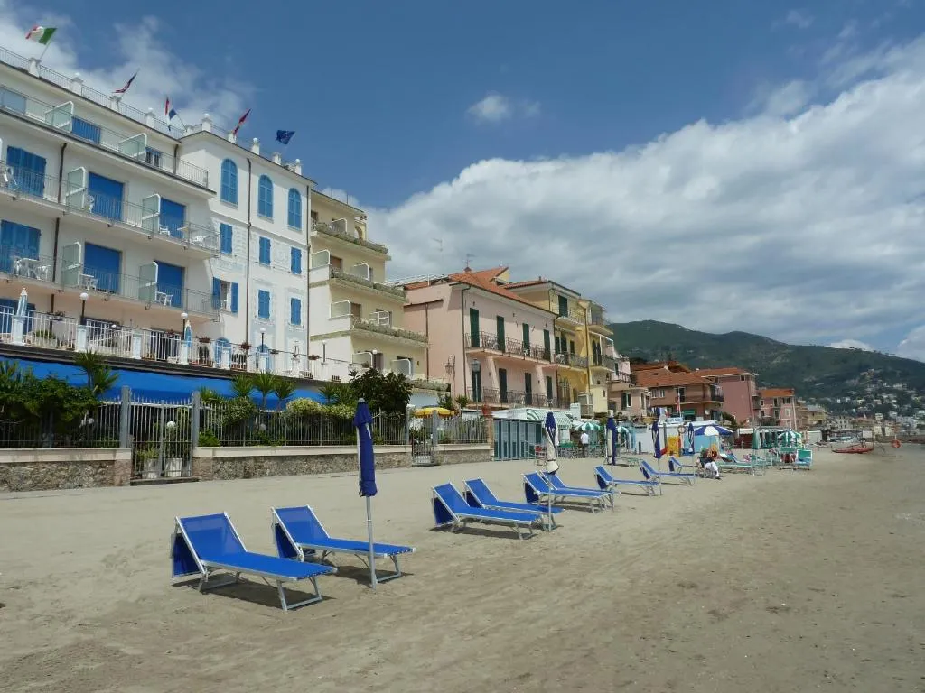 Spiaggia sabbiosa con lettini blu davanti all'hotel bianco con persiane azzurre e bandiere