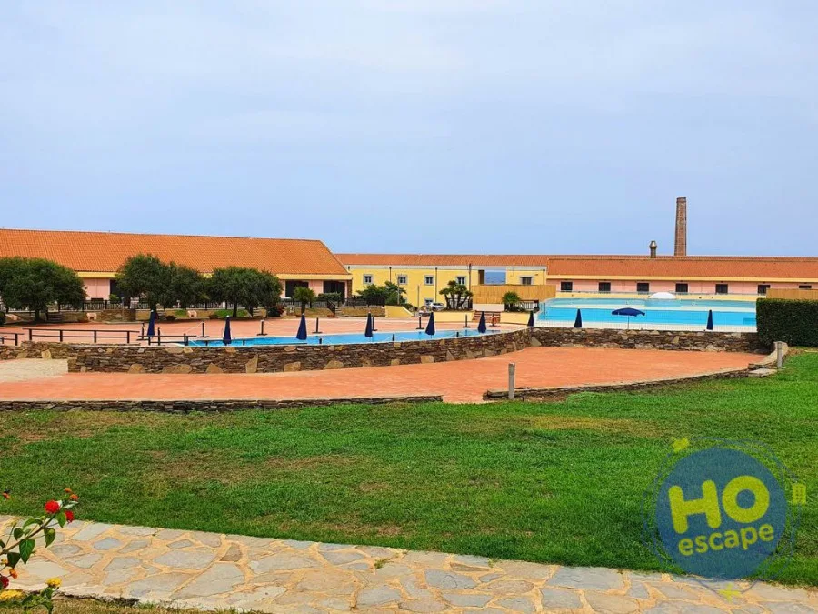 Vista del resort con piscine esterne, edifici color pastello e prato verde con vialetto in pietra