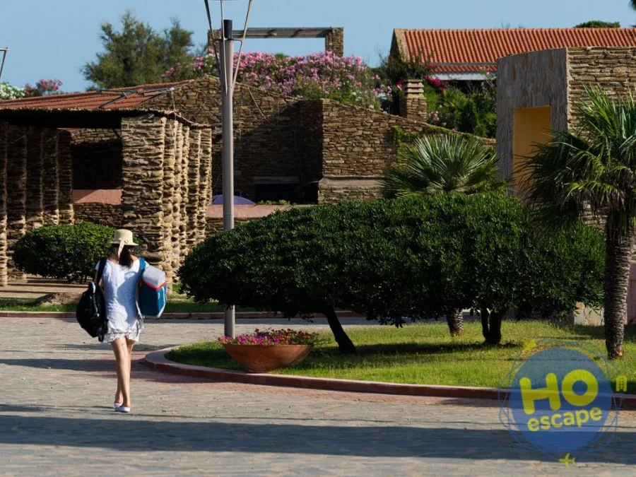 Ospite con cappello cammina lungo il vialetto del resort tra edifici in pietra e giardini curati