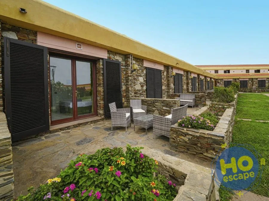 Patio esterno in pietra con poltrone in rattan e aiuole fiorite davanti alle camere dell'hotel