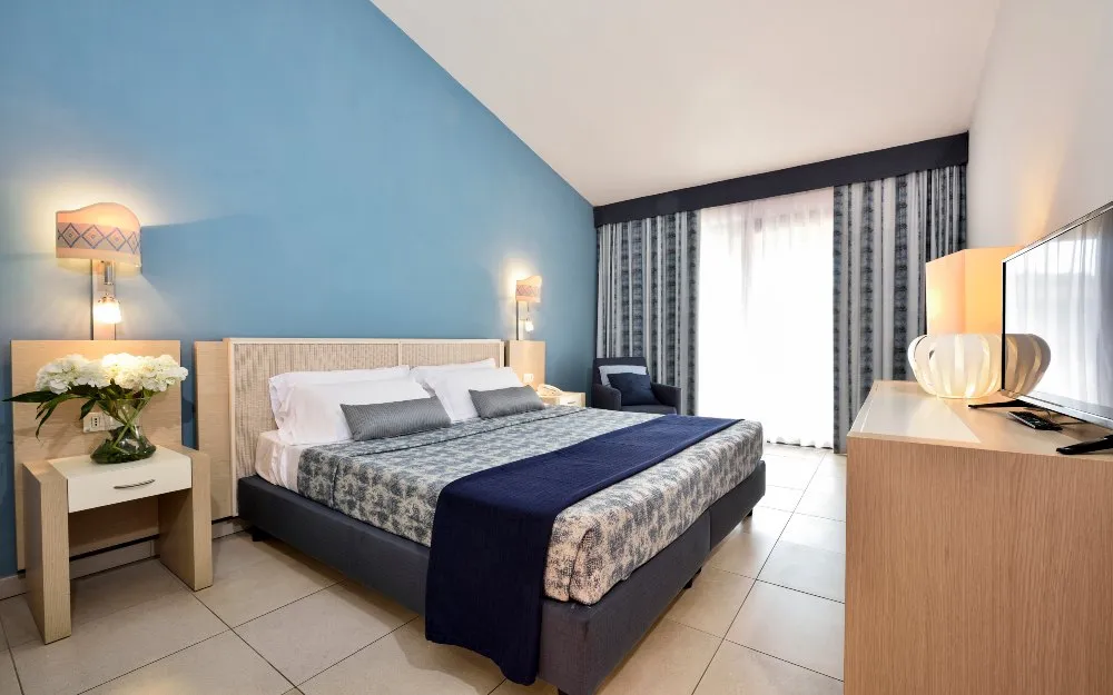 Camera matrimoniale con parete azzurra, letto, TV e ampia finestra con tende