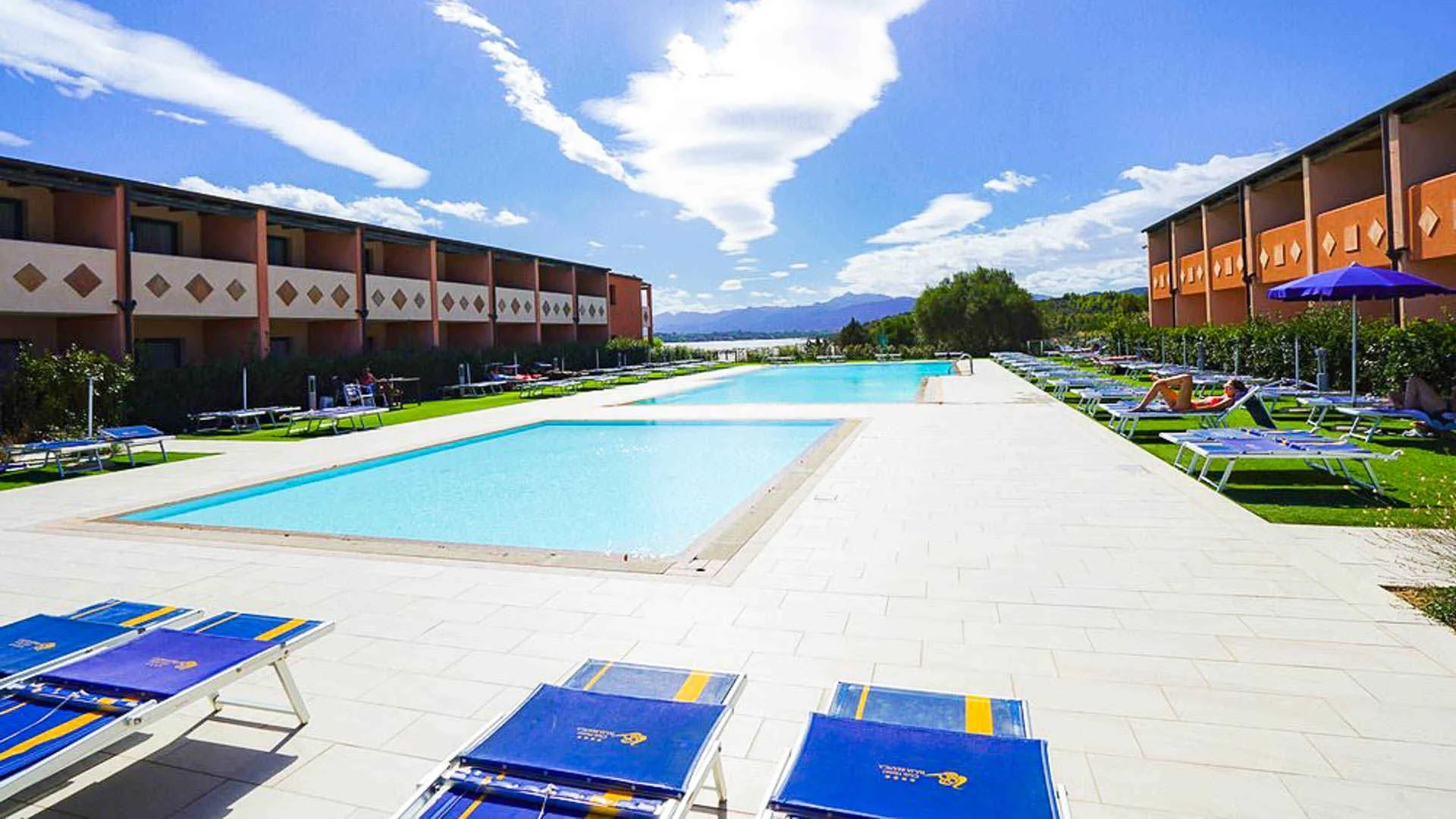 Due piscine rettangolari con pavimentazione chiara, lettini blu e vista sulla laguna