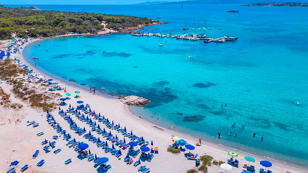 Veduta aerea di una spiaggia di sabbia chiara con ombrelloni blu e mare turchese cristallino con barche ormeggiate