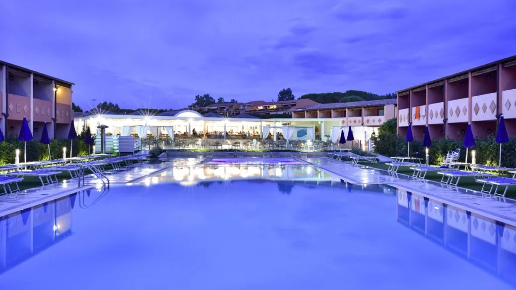 Piscina del resort illuminata al crepuscolo con riflessi viola e ristorante illuminato sullo sfondo