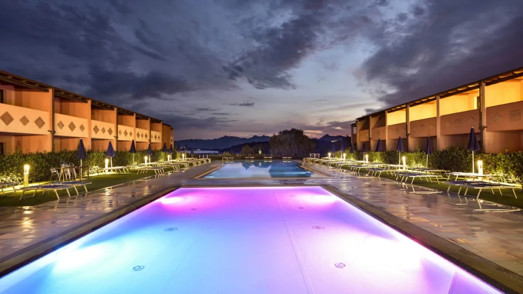Piscine illuminate al tramonto con giochi di luce rosa e blu tra gli edifici del resort