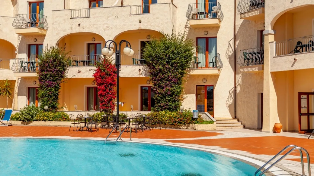 Bordo piscina con tavolini, sedie e piante fiorite davanti agli edifici