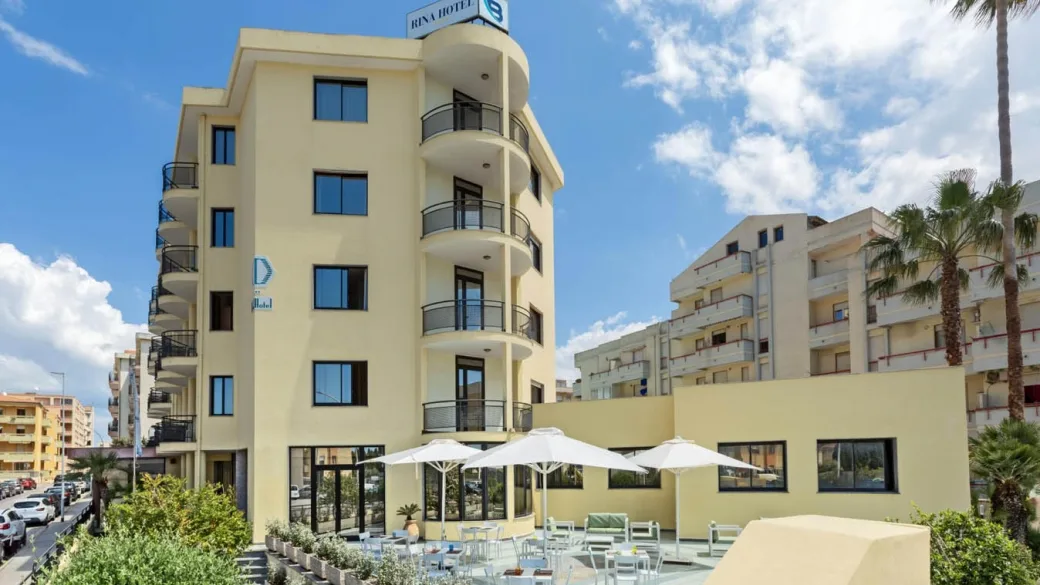 Vista esterna del Rina Hotel con balconi arrotondati, area relax e ombrelloni bianchi