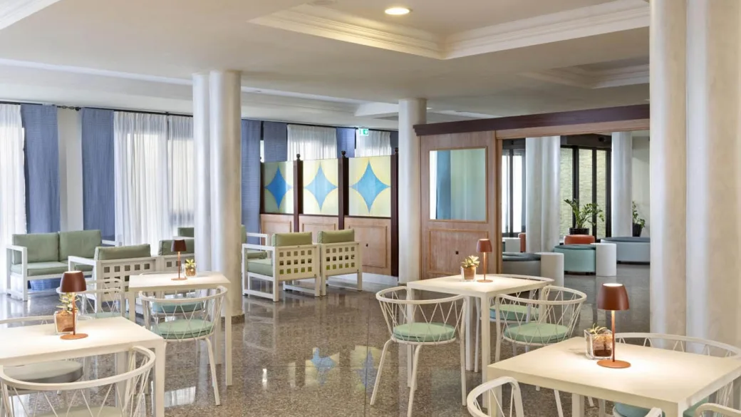 Sala comune dell'hotel con tavoli bianchi, poltrone verdi e lampade da tavolo
