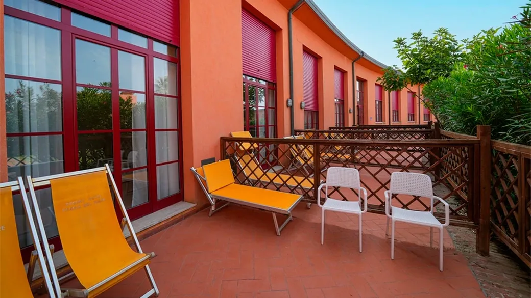 Terrazza esterna con lettini arancioni e sedie bianche lungo la facciata rossa dell'hotel