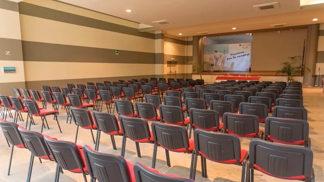 Sala conferenze con file di sedie rosse e nere e schermo per proiezioni