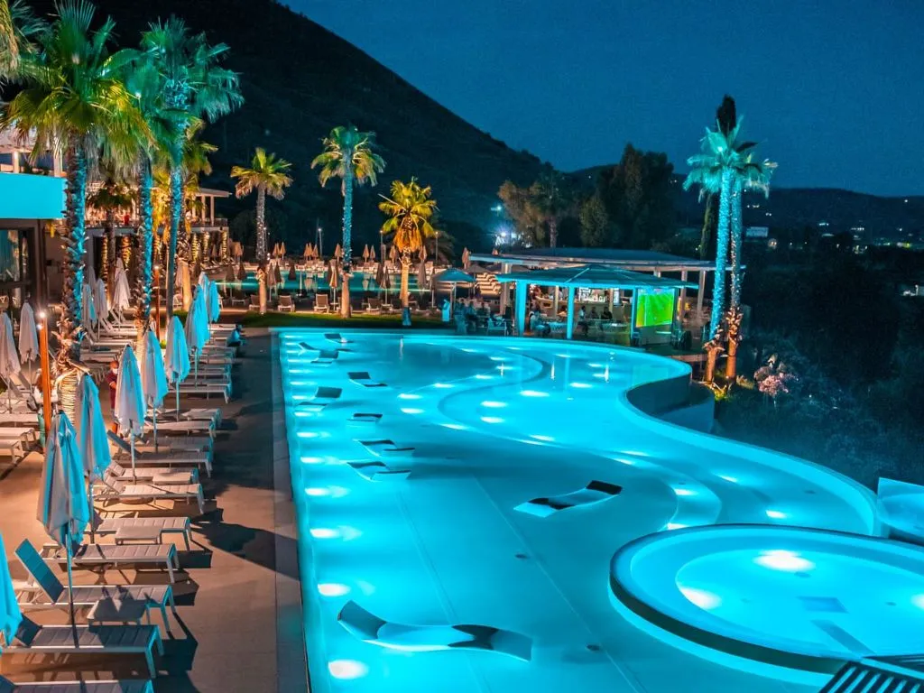 Piscina illuminata di sera con palme e area bar del resort
