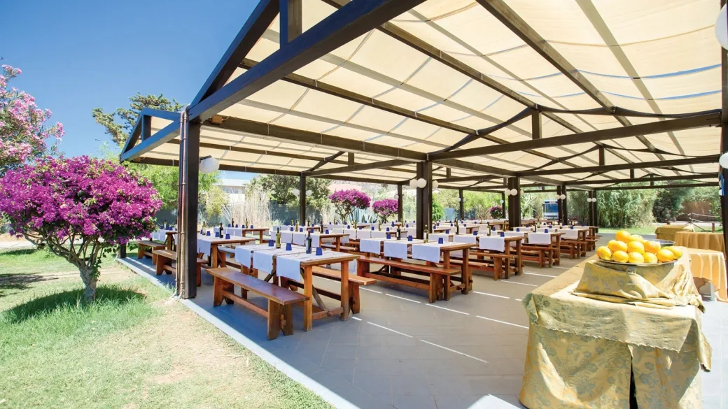 Area ristorante all'aperto con tavoli in legno sotto una pergola e piante fiorite