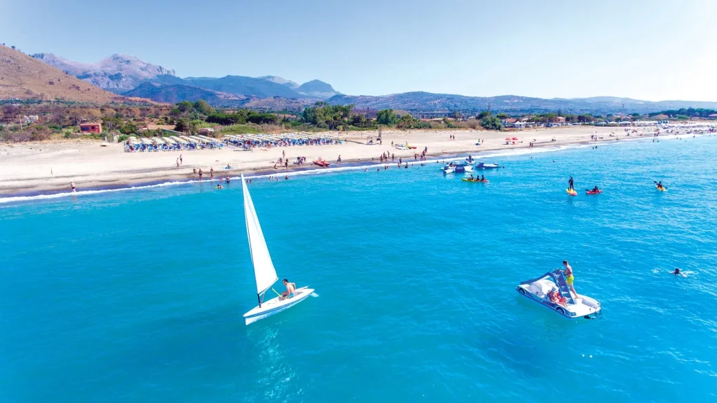 Barca a vela e pedalò nel mare turchese davanti alla spiaggia con montagne sullo sfondo