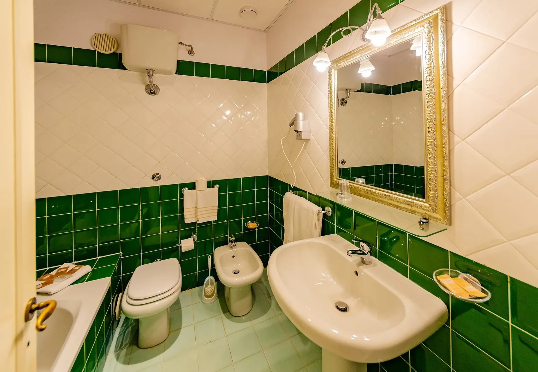 Bagno con piastrelle verdi e bianche, lavabo, bidet e specchio con cornice dorata