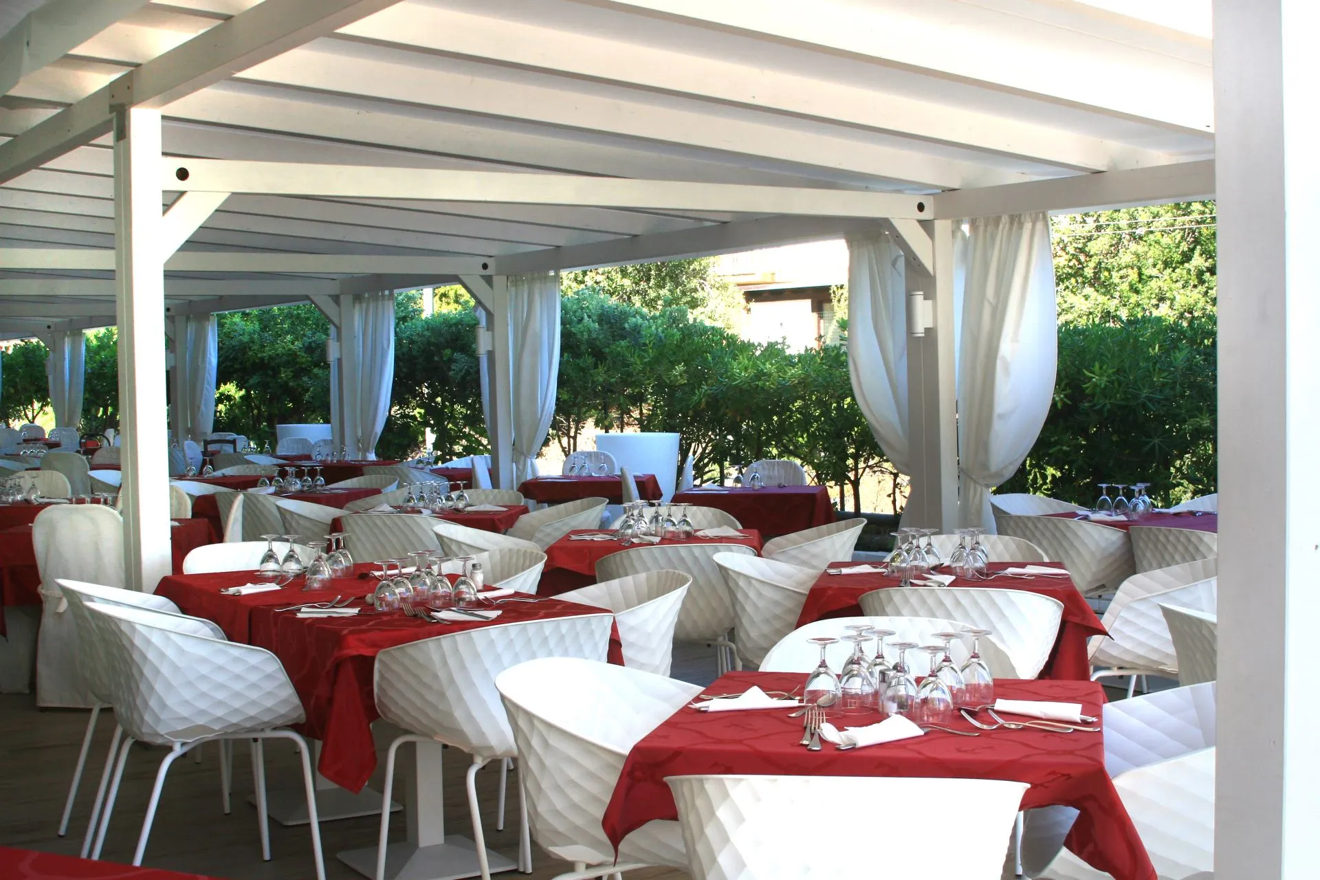 Ristorante esterno con tavoli tovagliati di rosso, sedie bianche e vegetazione