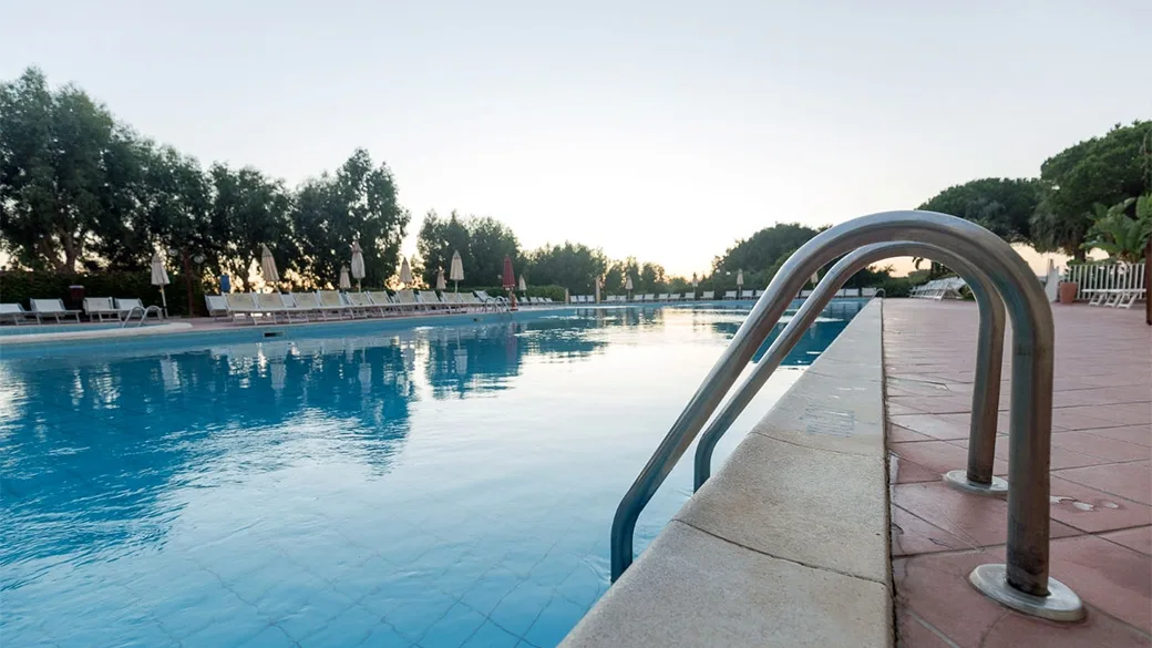 Bordo piscina al tramonto con scaletta in acciaio e lettini tra gli alberi
