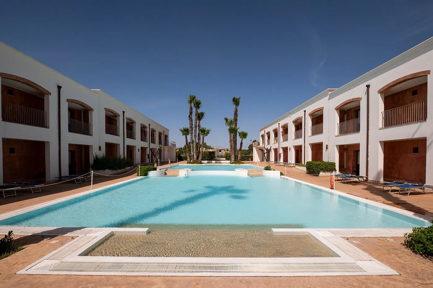Piscina rettangolare tra edifici bianchi con archi e palme sotto cielo azzurro