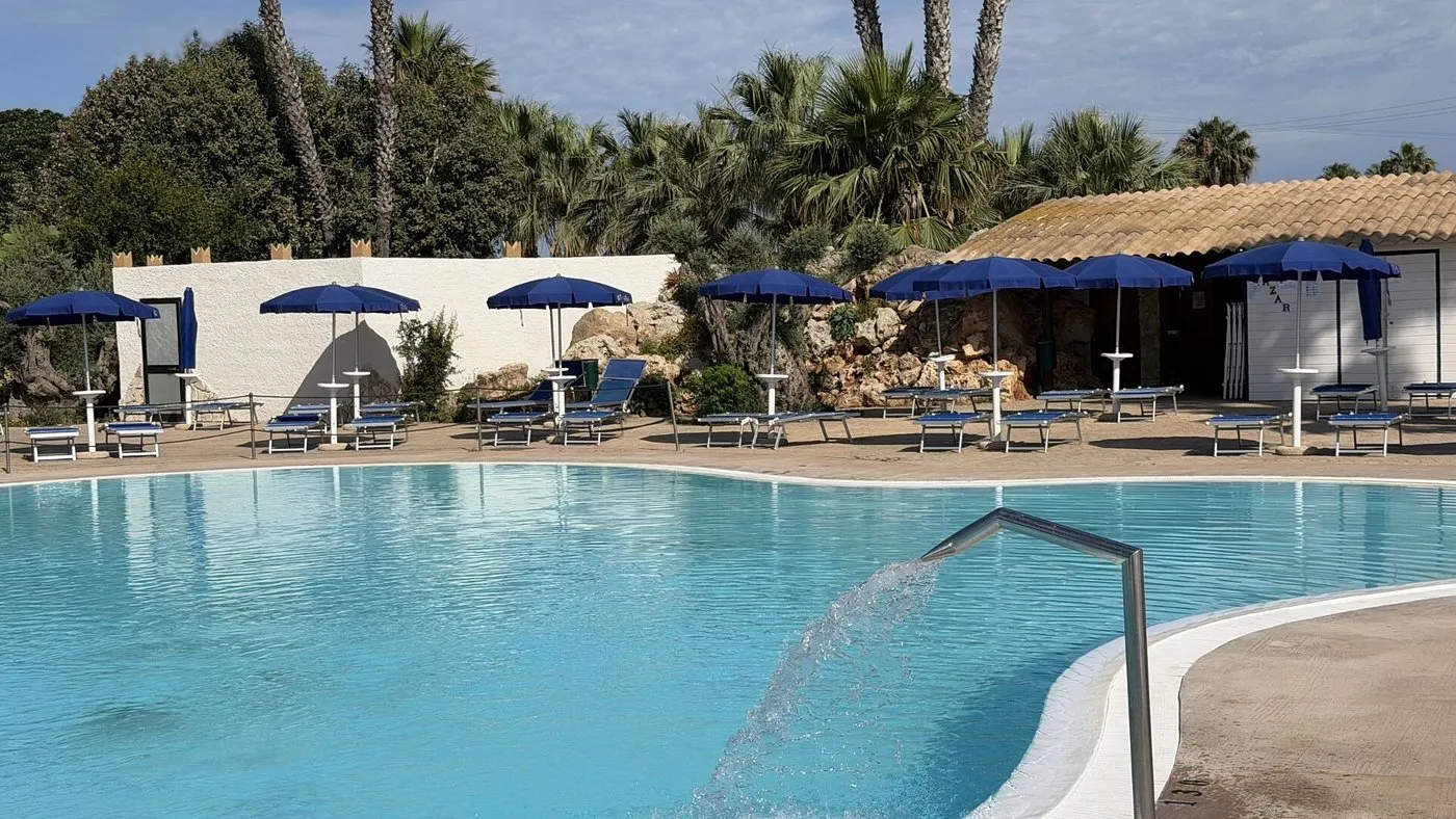 Dettaglio della piscina con getto d'acqua, lettini e ombrelloni blu tra le palme