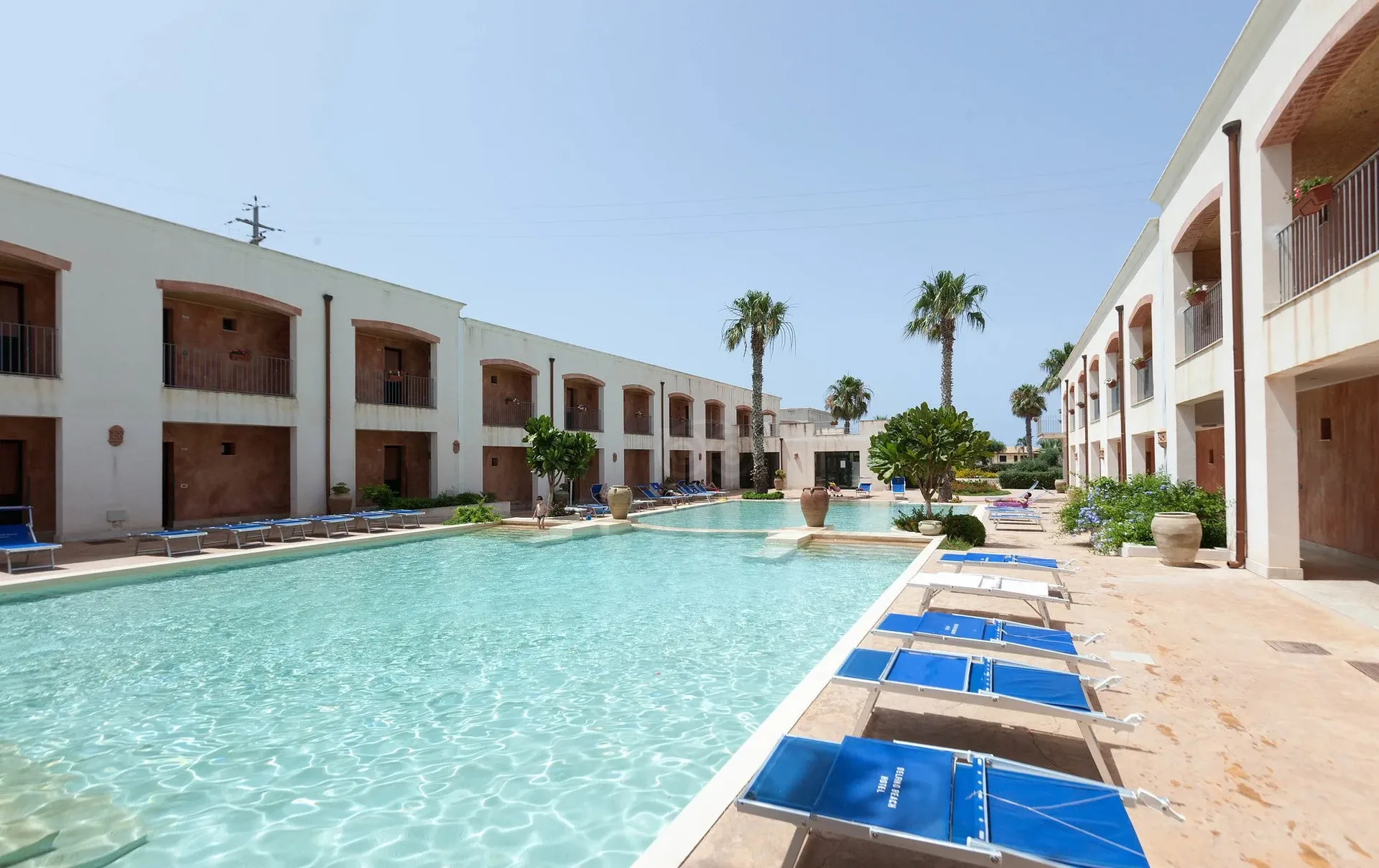 Lunga piscina tra edifici con archi e lettini blu a bordo vasca