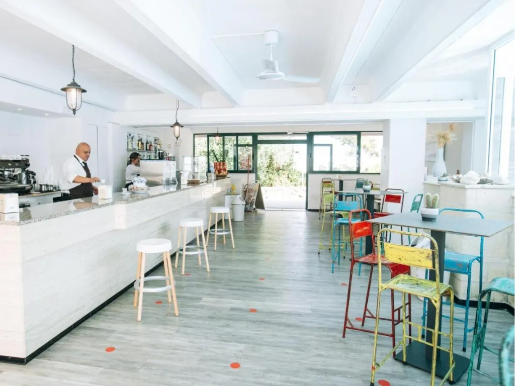 Bar interno luminoso con bancone bianco, sedie colorate e barista al lavoro